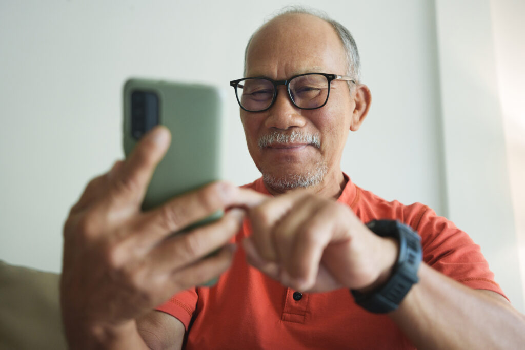 Man wearing eyeglasses and a red or orange shirt, smiling and using a smartphone.